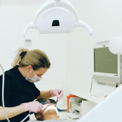 Focused woman in latex gloves and medical mask wearing black uniform holding medical instruments while treating teeth of male client in modern dental room